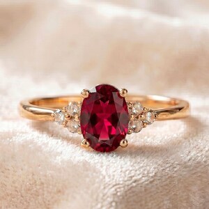 Könnte beinhalten: Ein goldener Ring mit einem zentralen, oval geschliffenen, tiefroten Edelstein, der von vier Krappen gehalten wird. Das Band ist auf beiden Seiten mit einer Gruppe von drei kleinen, klaren, rund geschliffenen Steinen verziert.