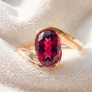 Op de afbeelding: Een gouden ring met een grote, ovaal geslepen, dieprode edelsteen in een bypass-stijl band. Het gepolijste goudmetaal buigt rond de gefacetteerde steen, wat zorgt voor een vloeiend ontwerp dat de levendige kleur van de edelsteen benadrukt.