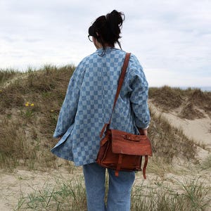 Pode incluir: Uma camisa de bot&otilde;es com padr&atilde;o xadrez azul-claro e branco, mangas compridas e colarinho, combinada com cal&ccedil;as de ganga azul-claro e uma mala de tiracolo em pele castanha com aba frontal, fivelas e al&ccedil;a de ombro ajust&aacute;vel.