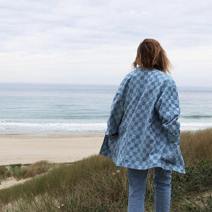 Pode incluir: Uma camisa de bot&otilde;es xadrez azul claro e branca com mangas compridas e gola, usada sobre cal&ccedil;as jeans azul claro. A camisa apresenta um padr&atilde;o quadrado repetitivo em todo o tecido e &eacute; mostrada a partir de uma perspectiva traseira.