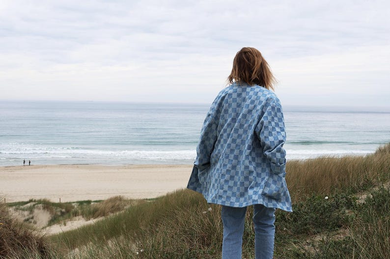 Pode incluir: Uma camisa de bot&otilde;es xadrez azul claro e branca com mangas compridas e gola, usada sobre cal&ccedil;as jeans azul claro. A camisa apresenta um padr&atilde;o quadrado repetitivo em todo o tecido e &eacute; mostrada a partir de uma perspectiva traseira.