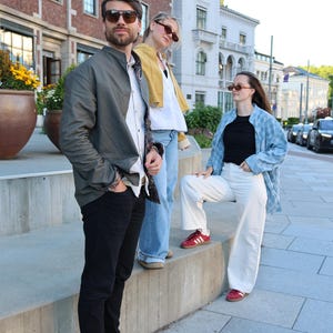 Pode incluir: Tr&ecirc;s pessoas vestindo roupas casuais: uma com jaqueta bomber verde-oliva, camisa branca e cal&ccedil;a jeans preta; outra com jaqueta curta amarela, camisa branca e cal&ccedil;a jeans azul; e uma terceira com camisa azul estampada, blusa preta e cal&ccedil;a branca.