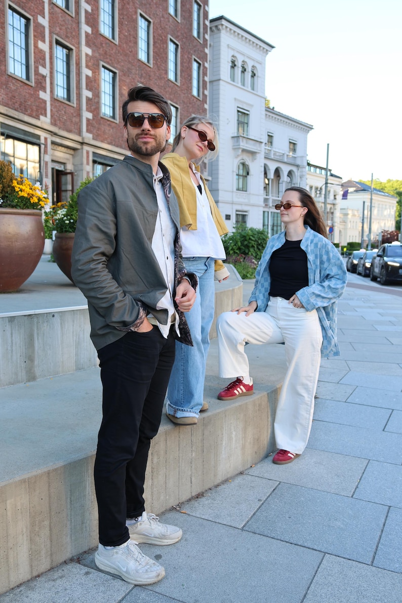 Pode incluir: Tr&ecirc;s pessoas vestindo roupas casuais: uma com jaqueta bomber verde-oliva, camisa branca e cal&ccedil;a jeans preta; outra com jaqueta curta amarela, camisa branca e cal&ccedil;a jeans azul; e uma terceira com camisa azul estampada, blusa preta e cal&ccedil;a branca.