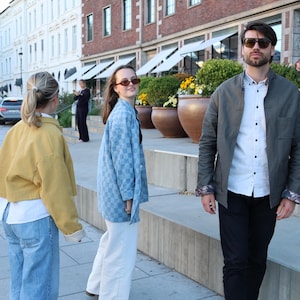 Pode incluir: Uma jaqueta amarela, uma camisa xadrez azul clara e uma jaqueta verde-oliva escura sobre uma camisa branca com cal&ccedil;as pretas. As pessoas usam &oacute;culos de sol enquanto est&atilde;o em uma pra&ccedil;a urbana pavimentada ao ar livre.