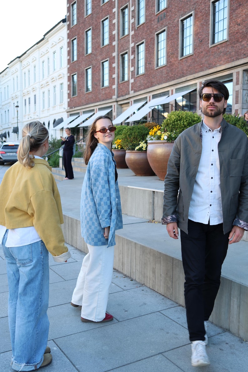 Pode incluir: Uma jaqueta amarela, uma camisa xadrez azul clara e uma jaqueta verde-oliva escura sobre uma camisa branca com cal&ccedil;as pretas. As pessoas usam &oacute;culos de sol enquanto est&atilde;o em uma pra&ccedil;a urbana pavimentada ao ar livre.
