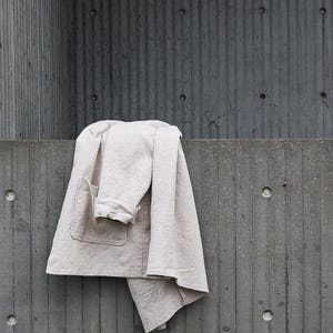 Pode incluir: Um casaco bege claro de mangas compridas com um bolso de remendo vis&iacute;vel na frente. A pe&ccedil;a apresenta uma superf&iacute;cie de tecido texturizado, uma gola dobrada e uma bainha reta, drapeada sobre uma superf&iacute;cie plana.
