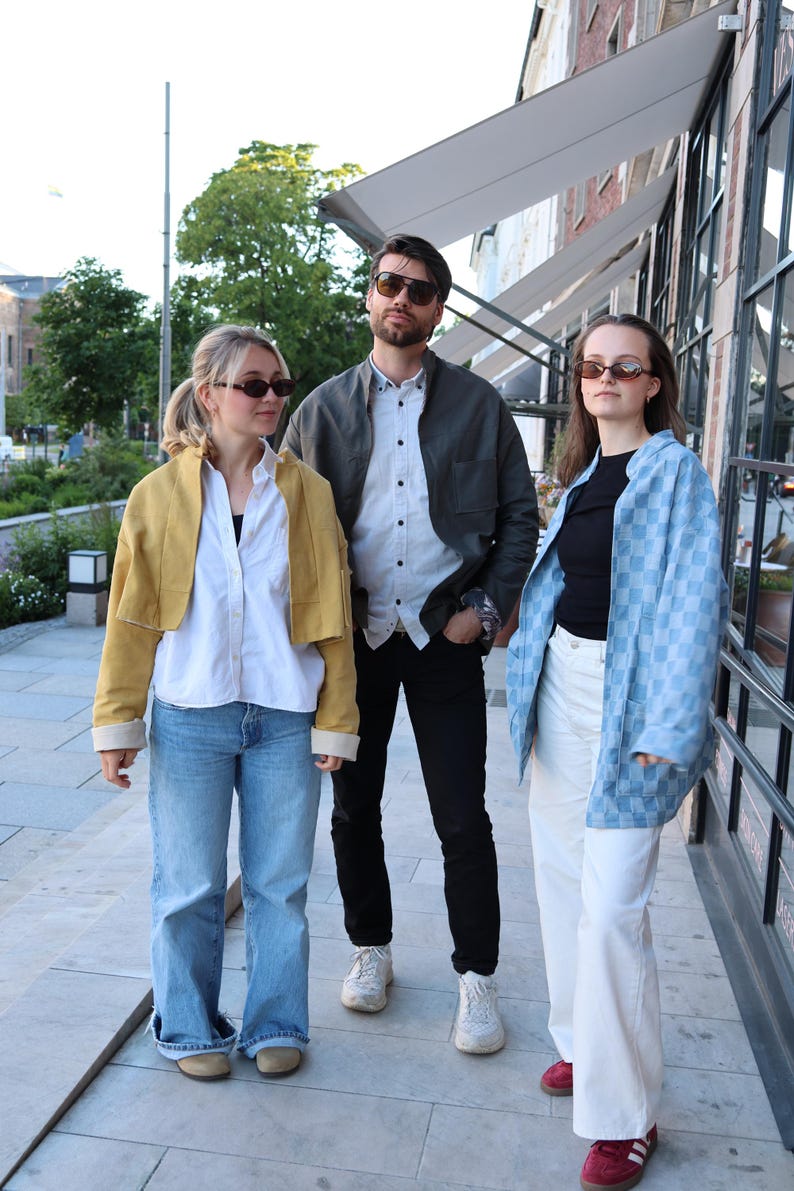 Pode incluir: Tr&ecirc;s pessoas vestindo roupas casuais. A pessoa &agrave; esquerda usa uma jaqueta curta amarelo-mostarda, camisa branca e cal&ccedil;a jeans azul. A pessoa ao centro usa jaqueta cinza-escura, camisa branca e cal&ccedil;a preta. A pessoa &agrave; direita usa jaqueta xadrez azul-clara, blusa preta e cal&ccedil;a branca.