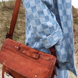 Pode incluir: Uma bolsa carteiro de couro marrom com uma al&ccedil;a longa, rebites de lat&atilde;o e uma aba, usada sobre uma camisa de manga comprida xadrez azul claro e branca e jeans azul claro. A bolsa apresenta costuras vis&iacute;veis e ferragens de metal.