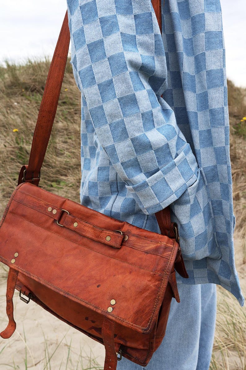 Pode incluir: Uma bolsa carteiro de couro marrom com uma al&ccedil;a longa, rebites de lat&atilde;o e uma aba, usada sobre uma camisa de manga comprida xadrez azul claro e branca e jeans azul claro. A bolsa apresenta costuras vis&iacute;veis e ferragens de metal.