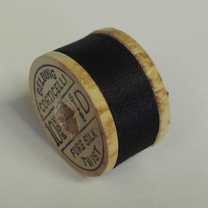 May include: A vintage wooden spool of black thread. The spool has a light brown wooden core with black thread wrapped around it. The label reads "Belding Corticelli" and "Pure Silk Twist".