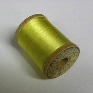 May include: A wooden spool of bright yellow thread. The spool features a circular label on the end with the text "GUARANTEED PURE SILK", "MADE IN THE U.S.A.", and "SHADE 957" printed in black ink around the central hole of the wooden spool.