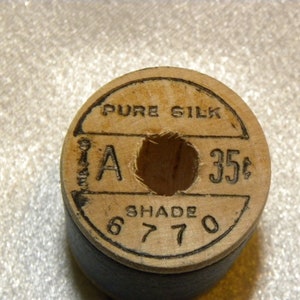 K&ouml;nnte beinhalten: Eine Holzspule mit reinem Seidenfaden und einem schwarzen Etikett. Das Etikett tr&auml;gt die Aufschrift "Pure Silk" und "35c" mit einem Buchstaben "A" und der Farbnummer "6770".