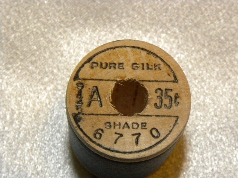K&ouml;nnte beinhalten: Eine Holzspule mit reinem Seidenfaden und einem schwarzen Etikett. Das Etikett tr&auml;gt die Aufschrift "Pure Silk" und "35c" mit einem Buchstaben "A" und der Farbnummer "6770".