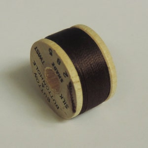 May include: A spool of dark brown silk thread for buttonholes. The wooden spool has the text "BUTTON HOLE SILK TWIST" and "SHADE 2 8". The thread is tightly wound around the spool, ready for sewing projects.