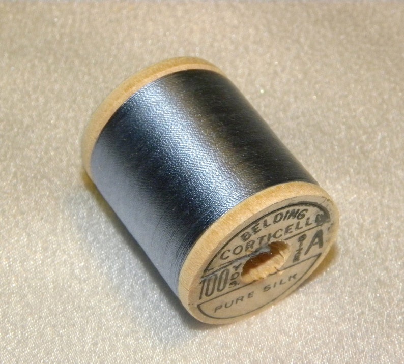 K&ouml;nnte beinhalten: Eine Spule blauen Seidenfadens mit einem Holzetikett mit der Aufschrift "Belding Corticelli 100% Reine Seide".