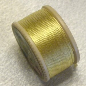 May include: A spool of yellow thread, tightly wound around a light beige plastic core. The thread has a slight sheen, and the spool is angled, showing the side and top. Ideal for sewing and crafting projects.