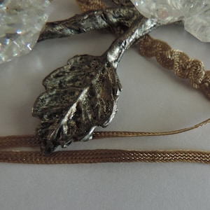 May include: A silver leaf pendant with a textured surface, attached to a brown cord. The pendant is surrounded by clear quartz crystals.