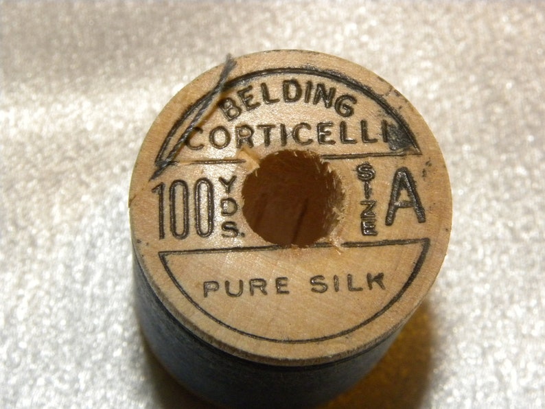 K&ouml;nnte beinhalten: Eine Holzspule mit schwarzem Garn mit der Aufschrift "Belding Corticelli 100 Yds. Size A Pure Silk"