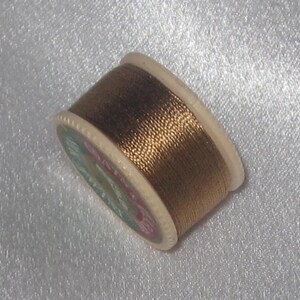 May include: A spool of metallic gold thread wound around a white plastic bobbin. The bobbin has a label with the text "DMC Satin" printed on it.