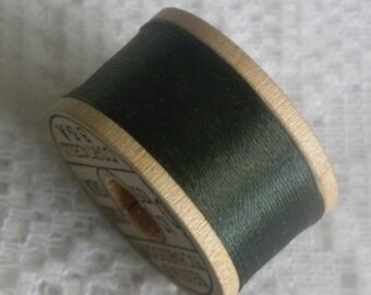 May include: A spool of dark green thread wound around a wooden bobbin. The bobbin is labeled with text in black.