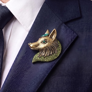Puede incluir: Un broche con forma de cabeza de zorro en metal dorado envejecido, con un ojo de cristal verde brillante, una pequeña piedra azul verdosa en la frente y un cuello texturizado incrustado con numerosas pequeñas piedras de estrás verdes brillantes.