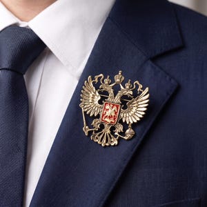 Pode incluir: Um broche de metal dourado com o emblema de uma águia de duas cabeças e um escudo central vermelho exibindo uma figura branca a cavalo. A águia é retratada com asas abertas, três coroas e segura um cetro e um orbe nas suas garras.