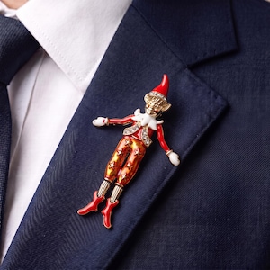 May include: A gold-tone metal brooch shaped like an elf, featuring red enamel accents on the hat, sleeves, and boots. The elf wears orange trousers with gold star patterns, a white collar, and clear rhinestones on the hat and vest.