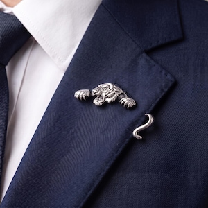 Op de afbeelding: Een zilverkleurige metalen broche in de vorm van een tijgerkop met uitgestrekte poten en een apart gebogen staartstuk, bevestigd op de revers van een donkerblauwe blazer gedragen over een wit overhemd met een donkerblauwe das.