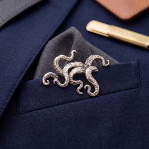 Puede incluir: Un broche de metal plateado con forma de pulpo con múltiples tentáculos curvos y texturizados que presentan pequeños detalles de ventosas circulares. El broche está colocado sobre un pañuelo de bolsillo azul oscuro en una chaqueta azul marino.