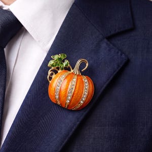 May include: A pumpkin-shaped brooch featuring bright orange enamel segments separated by rows of clear, sparkling rhinestones. The brooch includes a gold-toned stem and a green enameled leaf detail, pinned to the lapel of a dark navy blue suit jacket.
