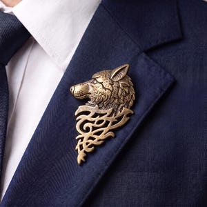 May include: A bronze-toned metal brooch shaped like a wolf head in profile, featuring detailed fur textures and a pointed ear, attached to a decorative filigree base with swirling cut-out patterns, pinned to the lapel of a dark navy blue blazer.