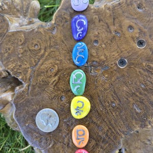 May include: A set of seven smooth, oval-shaped stones arranged in a vertical line, each featuring a unique hand-painted symbol in black. The stones are colored in a spectrum of light purple, blue, dark blue, green, yellow, orange, and red.
