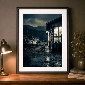 Op de afbeelding: Een ingelijste wandkunstprint met een donkere, regenachtige haven bij schemering. De afbeelding toont een houten gebouw met verlichte ramen, verschillende boten aangemeerd langs een natte pier en een dicht, donker bos onder een stormachtige lucht.