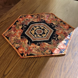 May include: A hexagonal fabric table topper featuring a seasonal autumn print with orange pumpkins, brown leaves, and corn stalks on a black background, centered with a geometric black and gold border pattern on a wooden dining table surface.