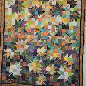 May include: A rectangular quilt featuring a patchwork design of small squares in various colors including green, orange, yellow, purple, and blue. The quilt displays several leaf-shaped patterns and is framed by a dark brown border.