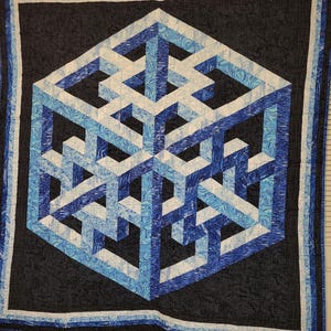 May include: A square quilt featuring a central geometric optical illusion of a three-dimensional cube. The design uses shades of blue, white, and black fabric with marbled patterns. The quilt is framed by a black border with a subtle grid texture.