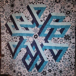 May include: A square quilt featuring a central geometric pattern of interlocking bands in shades of navy, teal, and light blue. The background is composed of a dense, repeating floral and snowflake-like print in varying tones of blue and white.