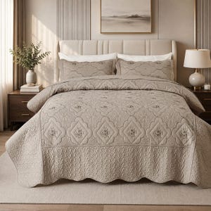 May include: A taupe-colored bedspread and two matching pillow shams featuring a quilted pattern with scalloped edges and embroidered floral motifs. The bedding set displays a repeating geometric design with intricate stitching across the fabric.