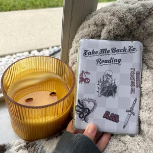 Może przedstawiać: Przezroczyste etui na telefon z napisem "Take Me Back To Reading" i różnymi gotyckimi naklejkami, w tym szkieletem, wiśniami i sztyletem. Obok etui stoi zapalona bursztynowa świeca.