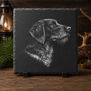 Könnte beinhalten: Eine quadratische schwarze Schieferplatte mit einem detaillierten, weiß gravierten Profilporträt eines Deutsch Drahthaar Hundes. Die Platte hat eine raue, natürliche Steinstruktur an den Kanten und wird von zwei kleinen schwarzen Metallständern gehalten.