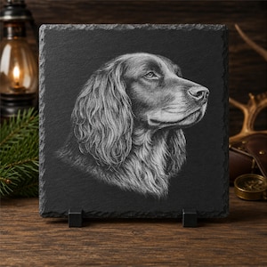 Könnte beinhalten: Eine quadratische schwarze Schieferplatte mit einem detaillierten, weiß gravierten Porträt eines Spaniels im Profil. Das Kunstwerk zeigt die langen, welligen Ohren und das strukturierte Fell des Hundes auf der natürlichen, dunklen Steinoberfläche.
