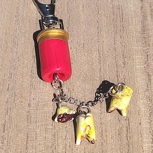 Può includere: Un portachiavi con un bossolo di fucile rosso e oro fissato a un moschettone in metallo argentato. Tre piccoli ciondoli a forma di dente dall'aspetto realistico sono sospesi alla base del bossolo da una catenella argentata, con smalto ingiallito.