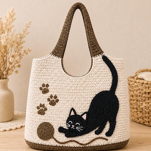 May include: A cream-colored crochet tote bag featuring a dark brown handle and base. The front displays an appliqued black cat stretching, a brown yarn ball, and three small brown paw prints. The cat has pink inner ears and white whiskers.