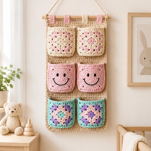 May include: A beige crochet wall hanging organizer featuring six pockets arranged in three rows. The top row has two cream and pink granny square pockets, the middle row has two pink pockets with black embroidered smiley faces, and the bottom row has two teal, purple, and pink granny square pockets.