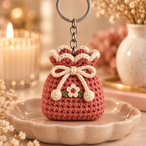 Puede incluir: Un pequeño llavero de bolsa de ganchillo en color rosa polvoriento con un cordón de ajuste color crema que termina en dos pompones redondos. La bolsa está decorada con una pequeña flor aplicada en el centro con pétalos blancos y una hoja verde.