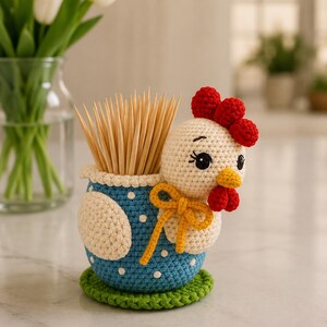 Peut inclure: Un porte-cure-dents en crochet en forme de poule avec un corps bleu à pois blancs, une tête crème, une crête et des barbillons rouges, et un bec jaune. La poule porte un ruban jaune noué autour du cou et repose sur une base verte.