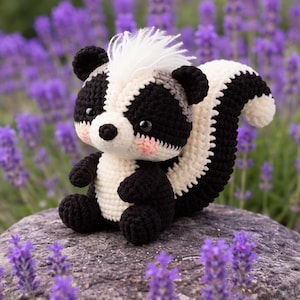 Op de afbeelding: Een gehaakt stinkdier knuffel gemaakt van zwart en crèmekleurig garen, met een pluizige witte kuif op het hoofd, een grote gestreepte staart en kleine zwarte ogen. Het knuffeldier heeft roze blosjes op de wangen en een gedetailleerd steekpatroon.