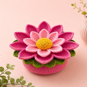 May include: A handcrafted crochet lotus flower container featuring a vibrant pink base and layered petals in shades of magenta and light pink. The center is a textured yellow circle, and the entire structure rests on a base of green leaves.