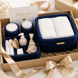 May include: A gift box containing navy blue crocheted baskets filled with baby care items including white nappies, cotton buds, a pump bottle, a wooden hairbrush, a small giraffe toy, and a card with the text Handmade with love.