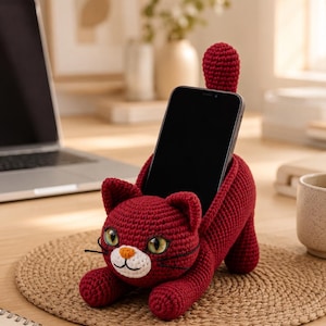 May include: A dark red crocheted cat phone holder featuring a textured yarn body, pointed ears, a white muzzle, and green eyes. The cat is in a crouching position with its tail upright, designed to securely hold a smartphone in its back slot.